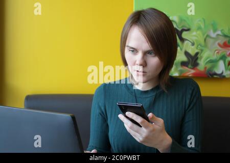 Una ragazza con un'espressione calma e attenta sul viso si siede a un computer portatile a un tavolo in un caffè su un divano marrone contro una parete gialla. Sta guardando un monitor del computer, tenendo uno smartphone, un telefono cellulare in mano. Foto Stock