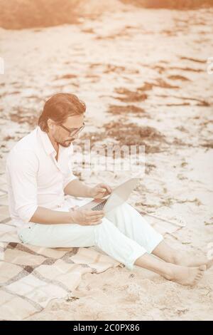 Uomo creativo che lavora con un computer portatile seduto su una spiaggia di sabbia. L'uomo caucasico di mezza età in abiti bianchi sta digitando sulla tastiera del computer, lavorando a distanza Foto Stock