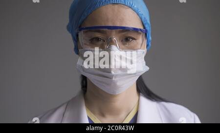 Medico in occhiali di sicurezza e maschera. Ritratto di donna asiatica in uniforme medica e occhiali di protezione guardando la macchina fotografica su sfondo grigio Foto Stock