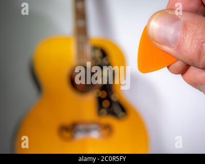 Primo piano di un uomo con un dito e un pollice che tiene un plettro di plastica arancione / Pick davanti a una chitarra acustica sfocata con finitura naturale. Foto Stock
