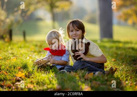 Bel ragazzo biondo di due anni, giocando con un aereo di legno e un orsacchiotto nel parco al tramonto, soleggiato giorno d'autunno Foto Stock