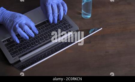 I guanti mani in mano umana funzionano su un computer portatile. Operatore medico che scrive sulla tastiera del computer situata su una scrivania di legno con un antisettico su di essa. Primo piano Foto Stock