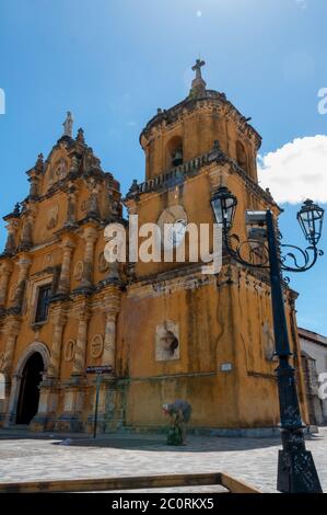 Vecchia laterna di fronte chiesa gialla con orologio bianco e una campana sulla cima sotto il cielo blu Foto Stock