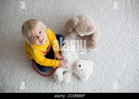 Bambino dolce, medico, che esamina il giocattolo dell'orso del bambino a casa, sfondo isolato Foto Stock