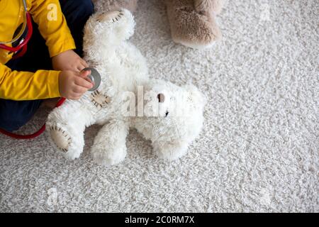 Bambino dolce, medico, che esamina il giocattolo dell'orso del bambino a casa, sfondo isolato Foto Stock