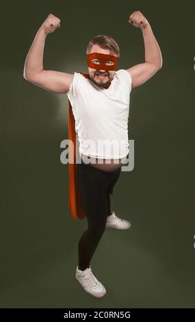 Forte supereroe che esprime potere. Uomo in maglietta bianca, maschera rossa e capo mostra bicipiti mentre si guarda la macchina fotografica. Isolato su verde erba cipollina Foto Stock