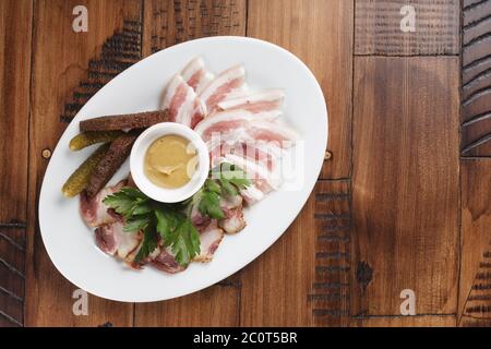 lardo affumicato e salato su un piatto bianco ovale. pancetta con salsa e verdure. antipasto di carne fredda. sfondo in legno. Foto Stock