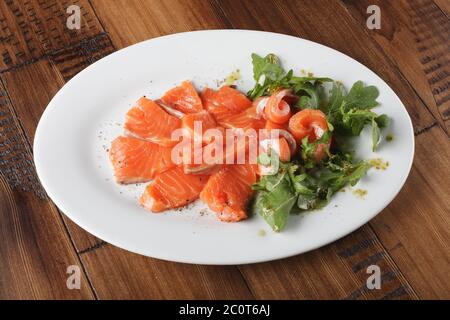 Trota affumicata con verdi su un piatto bianco. Sfondo di legno. Foto Stock