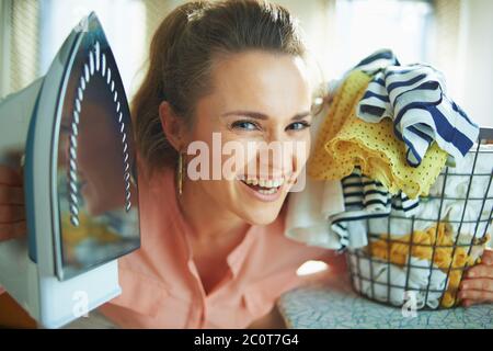 Ritratto di sorridente elegante casalinga di 40 anni in camicia rosa e pantaloni bianchi con ferro e cesto di vestiti lavati nella casa moderna in giornata di sole. Foto Stock