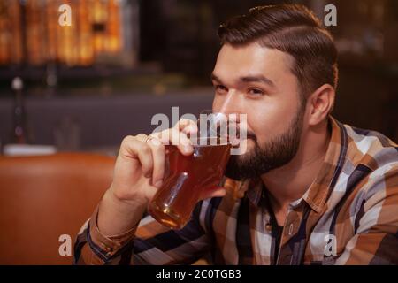 Primo piano di un uomo felice e rilassato che sorseggia una birra deliziosa, guardando via allegro. Un uomo bearded attraente che gusterete un bicchiere di birra al pub Foto Stock