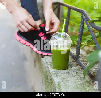 Coppa verde Detox frullato e donna allacciando scarpe da running prima dell'allenamento Foto Stock