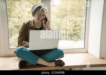 Donna freelance matura parla telefono cellulare che lavora computer portatile mentre si siede su misura-moda su silll di legno. Immagine colorata Foto Stock