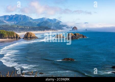 Vista panoramica verso Cannon Beach e Haystack Rock in una giornata di sole in Oregon. Dietro Haystack Rock arriva un muro di nebbia. Foto Stock