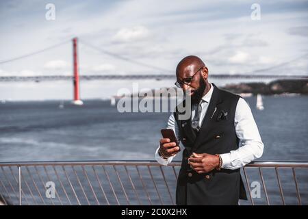 Un imprenditore africano serio ed elegante calvo in costume e occhiali sta usando il suo smartphone mentre si appoggia contro la scherma con un fiume Foto Stock