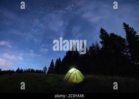 Magnifica notte estiva in montagna, magico cielo pieno di stelle sopra le alte cime degli abeti e tenda illuminata nel mezzo di prati di montagna. Spazio di copia. Concetto di viaggio Foto Stock