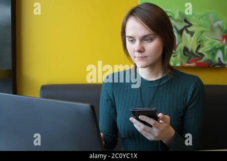 Una ragazza con un'espressione calma e attenta sul viso si siede a un computer portatile a un tavolo in un caffè su un divano marrone contro una parete gialla. Sta guardando un monitor del computer, tenendo uno smartphone, un telefono cellulare in mano. Foto Stock