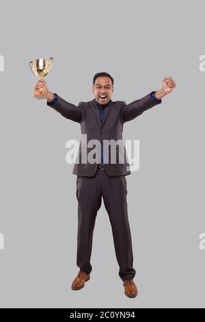 Uomo d'affari molto felice ed eccitato, alzando le armi, celebrando una vittoria o successo Holding Trophy. Firma del vincitore. Indiano uomo di affari isolato con Foto Stock