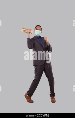 Uomo d'affari molto felice ed eccitato, alzando le armi, celebrando una vittoria o successo Holding Trophy indossare maschera medica. Firma del vincitore. Business indiano Foto Stock