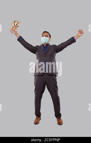 Uomo d'affari molto felice ed eccitato, alzando le armi, celebrando una vittoria o successo Holding Trophy indossare maschera medica. Firma del vincitore. Business indiano Foto Stock