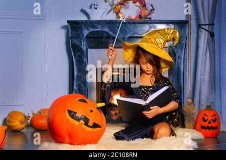 Halloween ritratto di mago ragazza sedersi con magic Stick cappello strega e incantesimo libro da camino Foto Stock