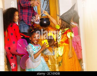 Ritratto di una ragazza sopra scena di stile di vita di Halloween con gruppo di bambini in costumi ricevono caramelle da donna che si trova vicino porta della casa insieme Foto Stock