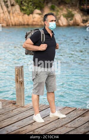 Uomo con maschera facciale, camicia nera e zaino in spalla è in vacanza e in background ha il mare e un resort turistico Foto Stock