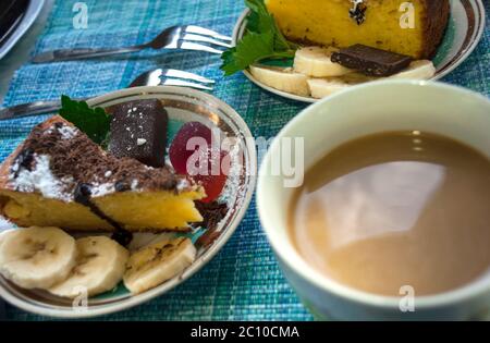 Porzione di torta con caramelle decorate con caramelle alla menta, toffee, banana e gelatina, zucchero in polvere e una tazza di caffè con latte su una tovaglia blu Foto Stock