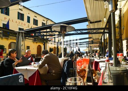 Persone e turisti seduti in un ristorante all'aperto nel centro storico sulla riva del Lago di Garda in una giornata di sole, Lazise, Verona, Veneto, Italia Foto Stock