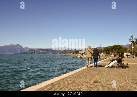 Vista panoramica sul Lago di Garda con persone e turisti che camminano e si siedono sulle panchine del lungolago in una giornata di sole, Lazise, Verona, Italia Foto Stock