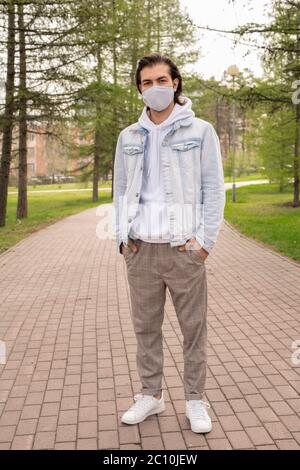 Bell'uomo in abito casual e maschera di stoffa che passeggiano nel parco della città durante la pandemia del coronavirus Foto Stock