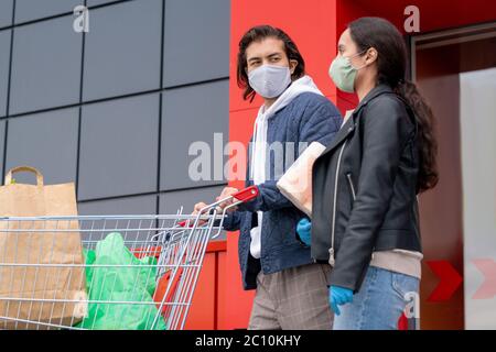 Giovane coppia in giacche e maschera di stoffa lasciando insieme supermercato dopo lo shopping, uomo che spinge il carrello Foto Stock