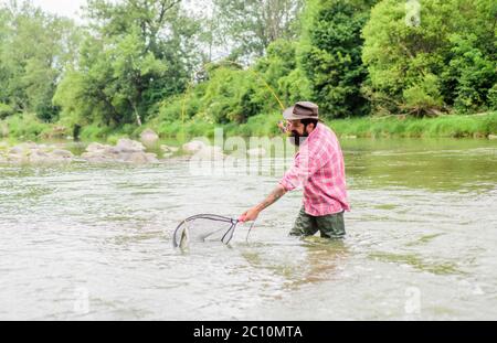 Hobby di pesca. pescatore brutale bearded che cattura il pesce di trota con la rete. Se pesce regolarmente sapete come ricompensare e calmante pesca è. La pesca è uno sport all'aperto accessibile con sedia a rotelle. Foto Stock