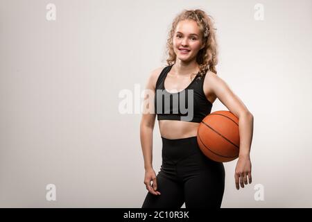 Felice giovane bionda giocatore di basket femminile in activewear nero che ti guarda con sorriso mentre tenendo la palla tra vita e braccio Foto Stock