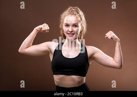 Forte atleta giovane attiva in tuta nera che fa gesto mostrando la sua potenza fisica mentre si trova in posizione isolata Foto Stock