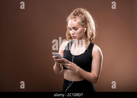 Giovane bionda femminile millennial in tuta nera e auricolari che scorrono attraverso la playlist nello smartphone, mentre si sceglie qualcosa da ascoltare Foto Stock