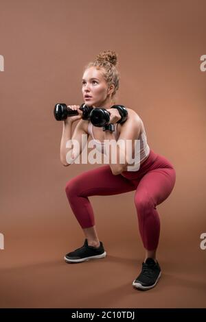 Giovane sportivo in tuta seduta su squadre e sollevamento di manubri durante un esercizio difficile Foto Stock
