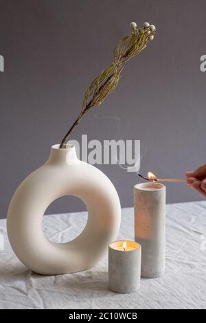 Mani di giovane donna che tiene la partita bruciante su una delle due candele aromatiche in bianco vetro di ceramica fatto a mano in piedi da vaso Foto Stock