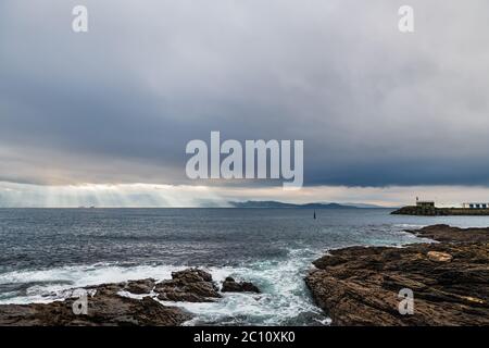 Scogliere e piccolo faro in una giornata invernale nuvolosa a Portonovo in Galizia, Spagna, con l'ingresso alla Ria de Pontevedra sullo sfondo. Foto Stock