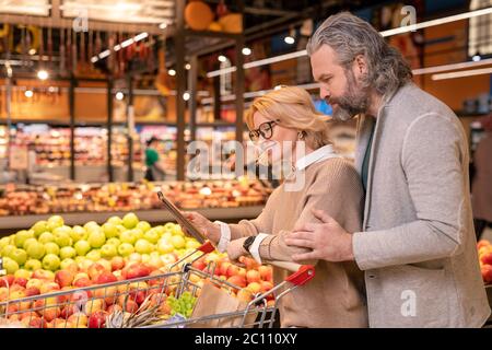 Felice giovane shopper donna che mostra la sua lista di acquisti marito in blocco note mentre entrambi camminando lungo esposizione con mele fresche Foto Stock