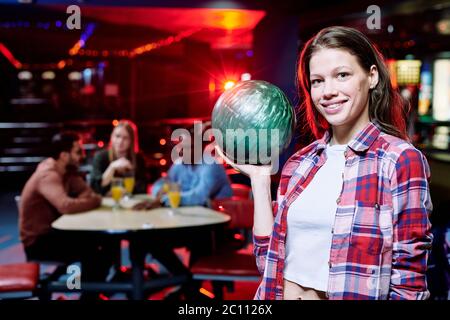 Bella ragazza sorridente in casualwear tenendo palla da bowling mentre si trova di fronte alla fotocamera sullo sfondo di gruppo di amici interculturali Foto Stock