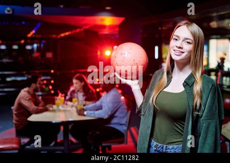 Felice giovane donna casual con lunghi capelli biondi che tengono la palla da bowling mentre gioca al centro ricreativo con i suoi amici sullo sfondo Foto Stock