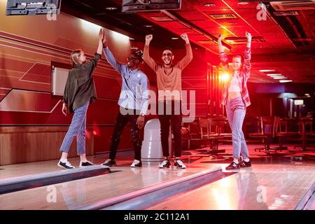 Giovane coppia interculturale eccitata dandosi a vicenda cinque alti mentre i loro amici allegri con le mani alzate in piedi dalla pista di bowling Foto Stock