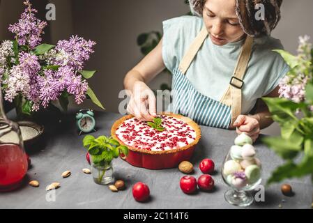 Cucina torta di mirtilli fatti in casa. Una pasticciera femminile sta tenendo una crostata di mirtillo preparata con panna acida bianca. Tavolo con ingredienti luminosi Foto Stock