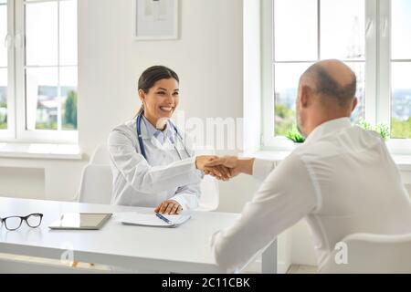 Donna medico ispanica sorridendo scuotendo le mani con un paziente anziano presso l'ufficio medico. Foto Stock