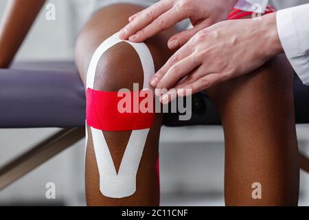 Fisioterapista applicando nastro di cinesiologia al paziente knee.Therapist che tratta giovane atleta afro-americana femminile. Nastro per cinesiologia. Post traumatico Foto Stock