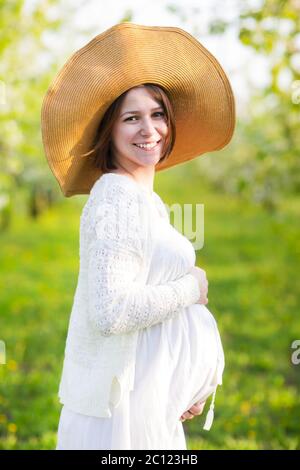 Donna incinta che indossa un cappello grande e un abito bianco in giardino fiorito Foto Stock