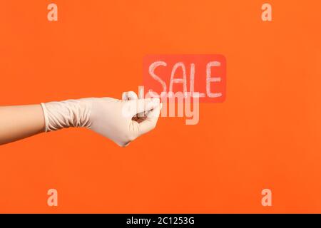 Profilo Vista laterale primo piano della mano umana in guanti chirurgici bianchi che tengono e mostrano etichetta vendita rossa. Interno, studio shot, isolato su sfondo arancione. Foto Stock