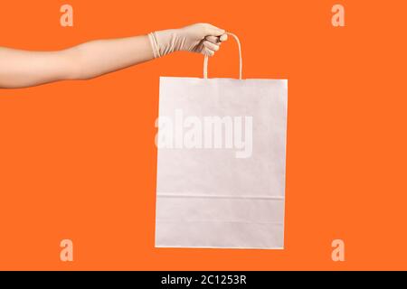 Profilo Vista laterale primo piano della mano umana in guanti chirurgici bianchi che tengono e mostrano la borsa da shopping bianca. Interno, studio shot, isolato su dorso arancione Foto Stock