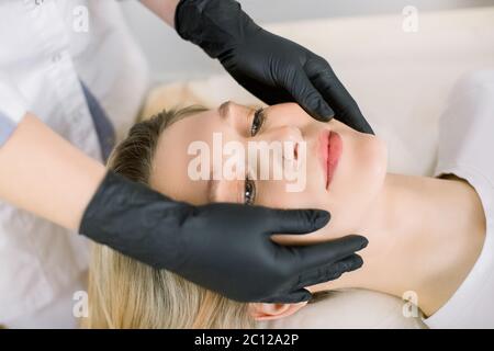 Vista laterale immagine ritagliata delle mani di donna cosmetologa che fa massaggio del viso a giovane donna sorridente sentirsi rilassato in salone di bellezza. Concetto di Foto Stock