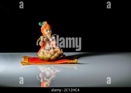 Una scultura isolata del dio indiano lord Krishna che gioca flauto seduto su un tappetino. Su un tavolo bianco riflettente con sfondo scuro Foto Stock